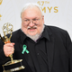 George R.R. Martin holds aloft an Emmy statue while wearing a black shirt and jacket.
